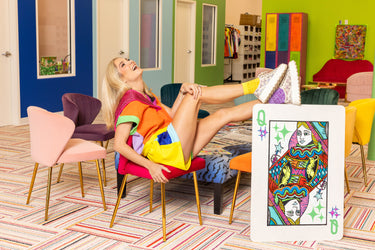 Company Founder, Jaime Glas Odom, is pictured in Queen of Sparkles headquarters in Baton Rouge, Louisiana. She is seated in a bright fuschia chair and is wearing a multi-colored sequin short set in bright shades of orange, yellow, lilac, and fuschia. The company logo is pictured next to here, which is a multi-colored rendition of a Queen from a deck of playing cards, reimagined to include sparkly diamonds, tigers, and magic wands. 
