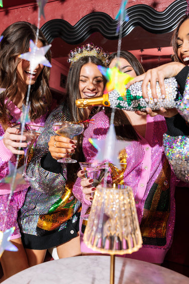 Several girls gather together as one of them pours them glasses of champagne. Each girl is dressed in sparkly party dresses in pink, black, and silver. 