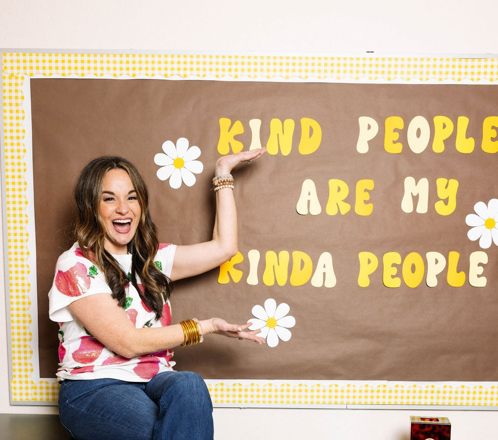 A brunette teacher in a white, sequined t-shirt with apple motif and blue flared jeans points to a sign in her classroom that reads "Kind people are my kinda people." The teacher is sitting on a table in her high school classroom.