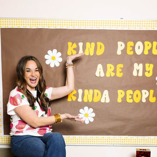A brunette teacher in a white, sequined t-shirt with apple motif and blue flared jeans points to a sign in her classroom that reads "Kind people are my kinda people." The teacher is sitting on a table in her high school classroom.